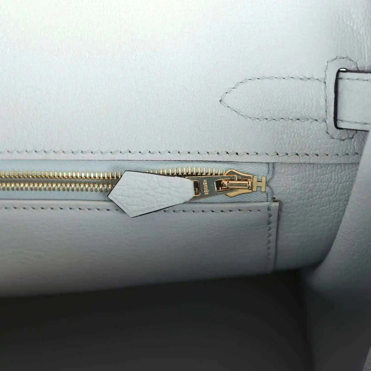 Pale blue leather handbag close-up of gold zipper pull and small tab near stitching