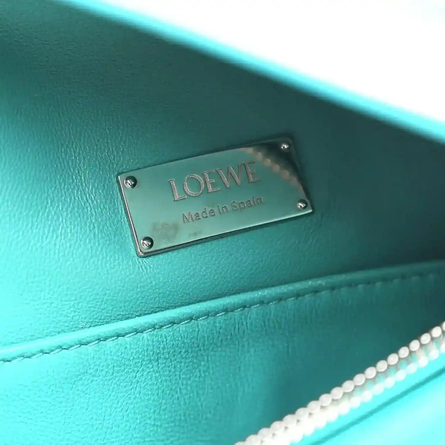 Teal-blue leather bag label plate reading LOEWE and Made in Spain.