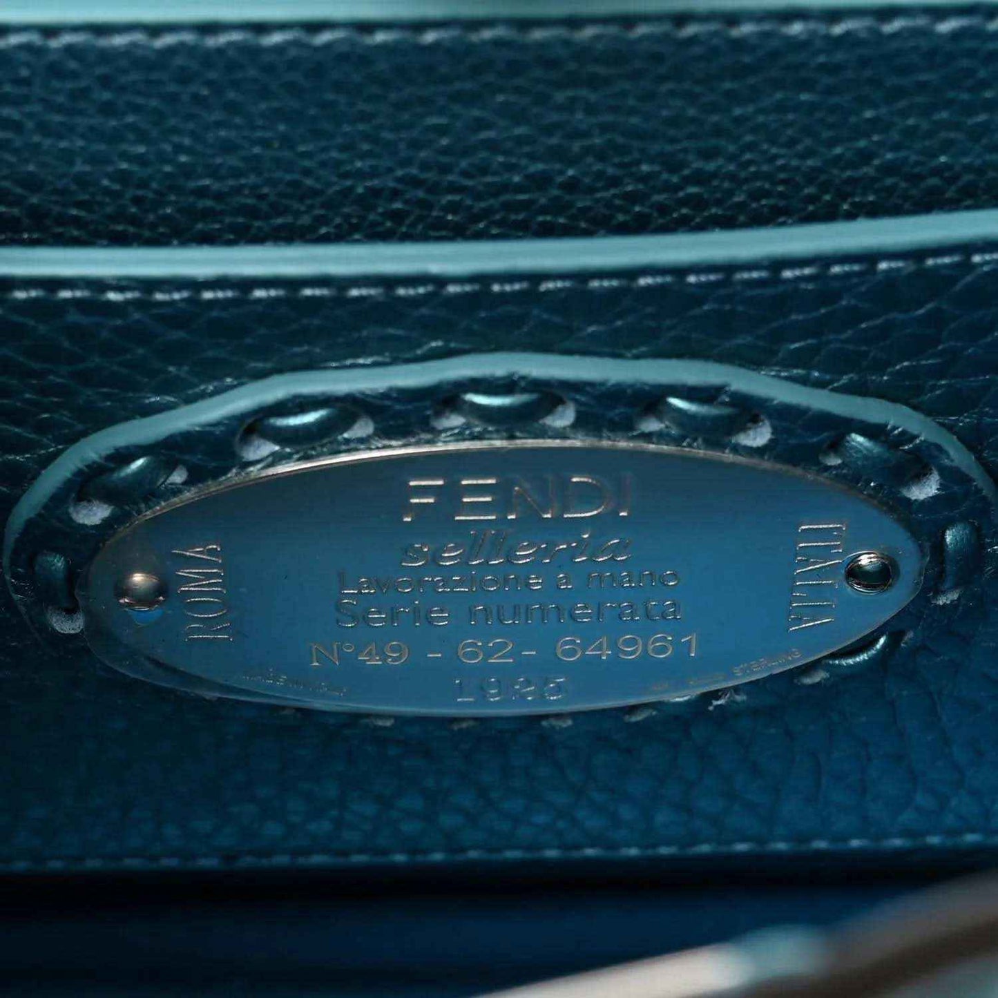Blue leather satchel tag with Fendi logo and serial text on front hardware.