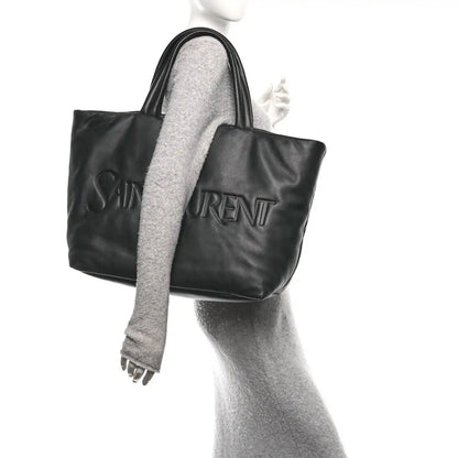 Black lambskin wide puffer tote with embossed logo, top handles, and side view.