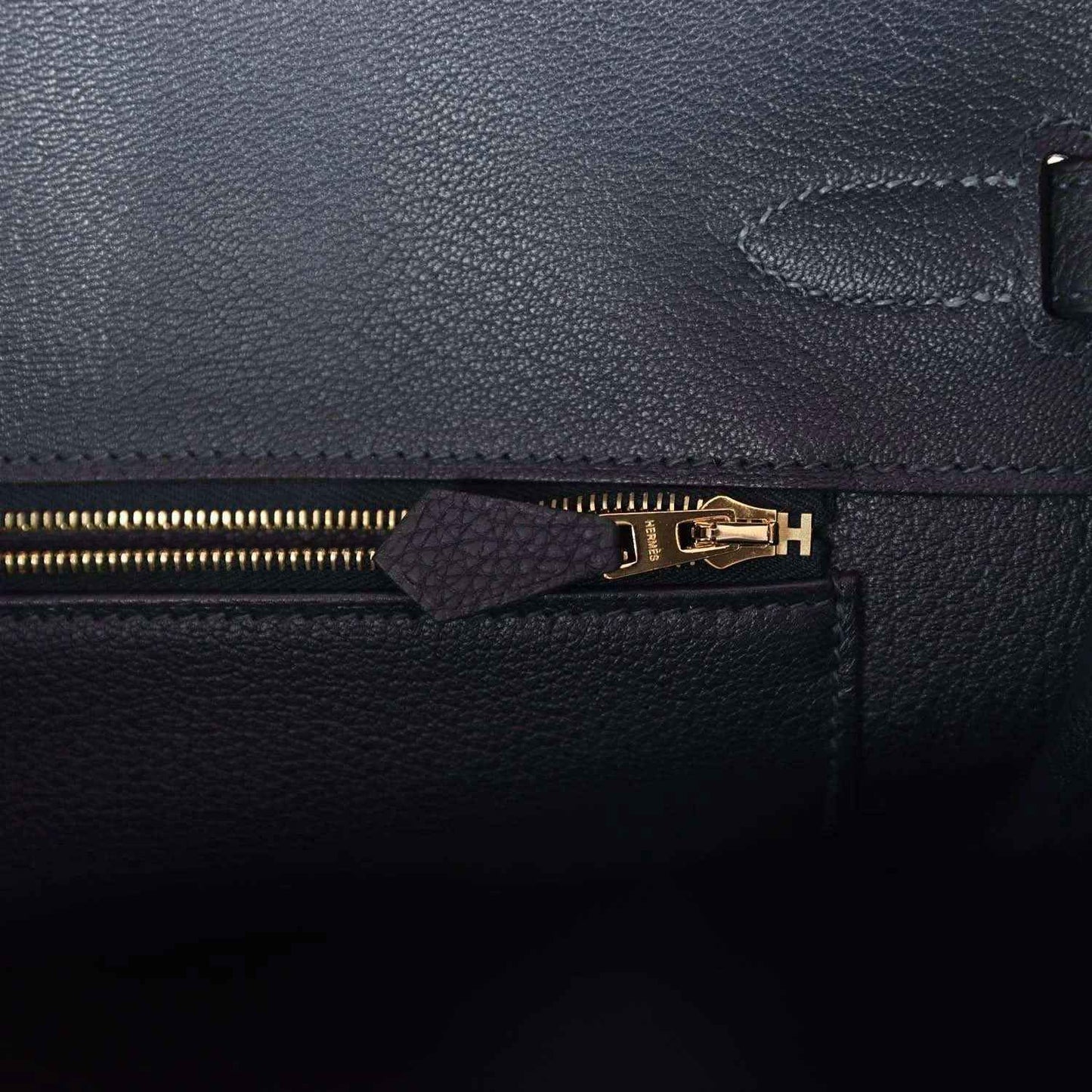 Leather handbag zipper close-up with gold teeth and small leather pull tab, dark color
