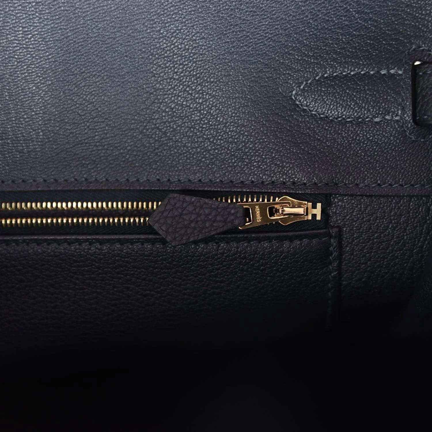 Leather handbag zipper close-up with gold teeth and small leather pull tab, dark color