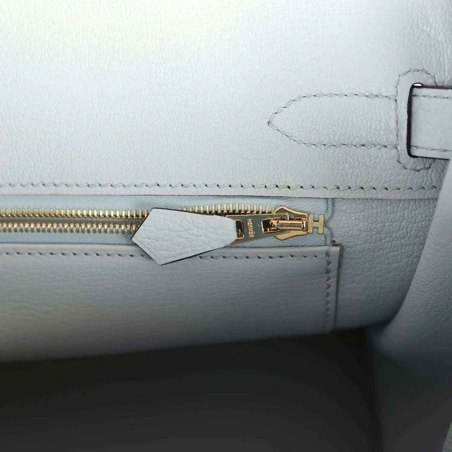 Pale blue leather handbag close-up of gold zipper pull and small tab near stitching