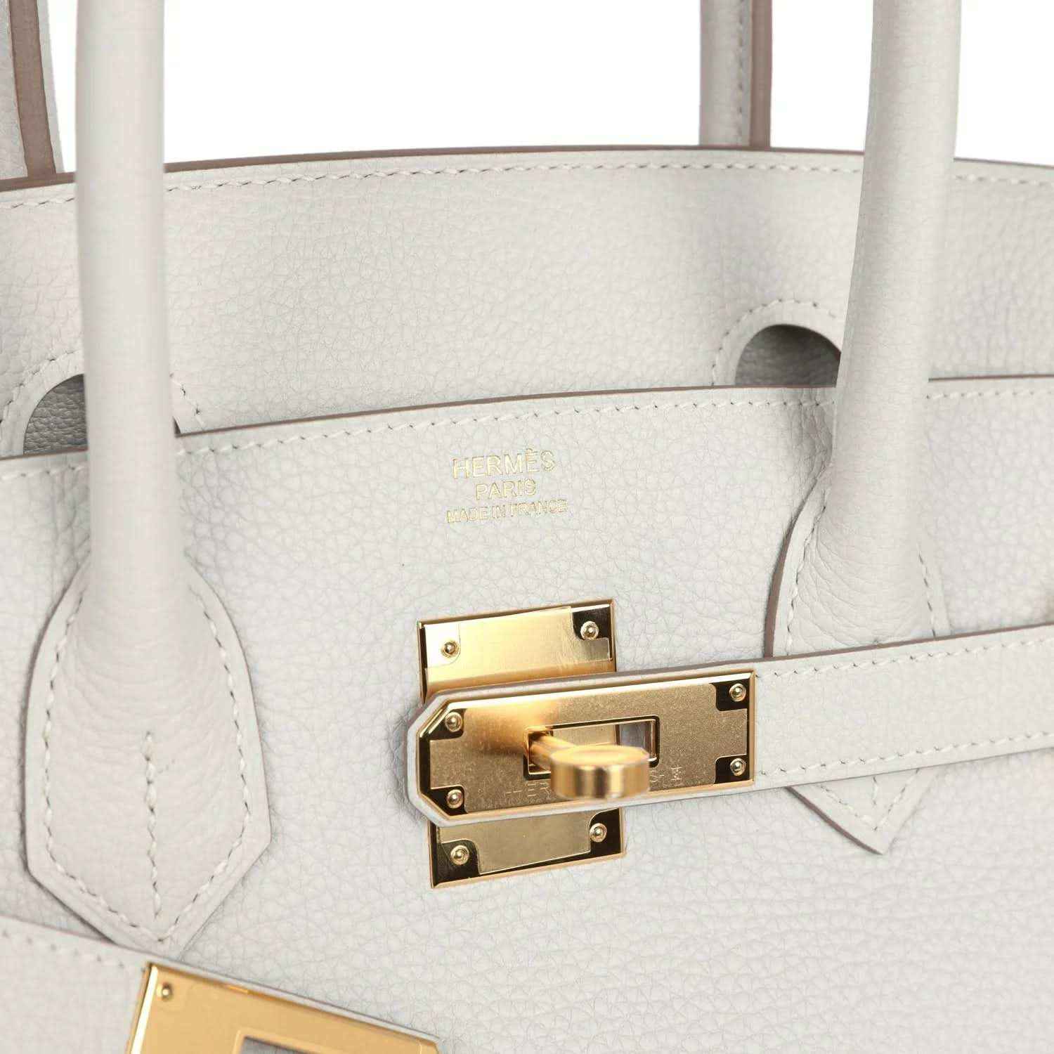 Light gray leather handbag side with gold turn-lock hardware and stitched leather trim