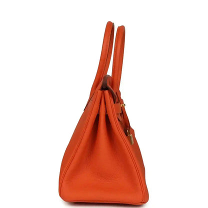 Orange leather handbag side view with two top handles and structured silhouette.