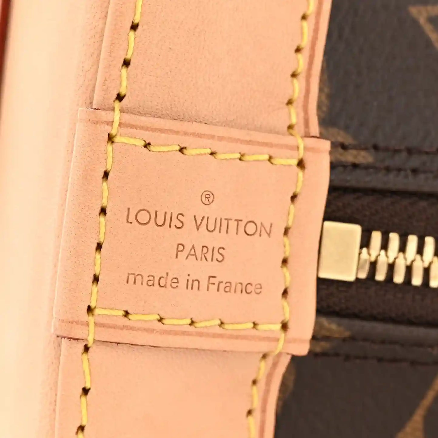 Tan leather label detail on monogram tote bag with stitched text and gold zipper edge