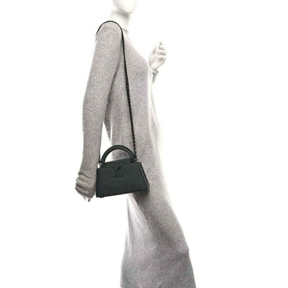 Mini black leather handbag with short handle and crossbody strap, held at side.
