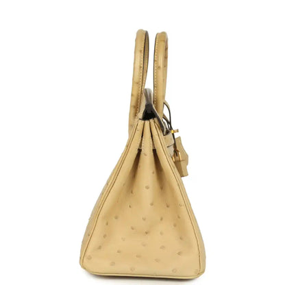 Tan ostrich leather handbag viewed from the side, with top handles and hanging tassel.