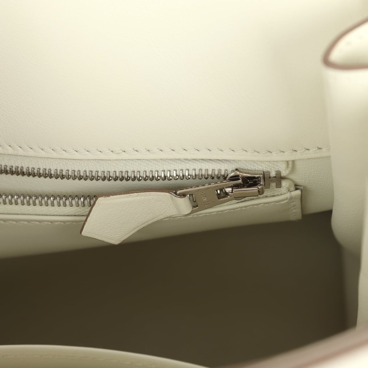 Cream leather handbag interior close-up of zipper track with metal pull tab.